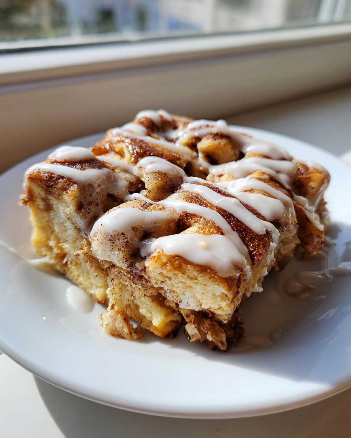 A close-up of a serving of Easy Sheet Pan Cinnamon Roll Pancake Bake drizzled with white icing on a white plate.