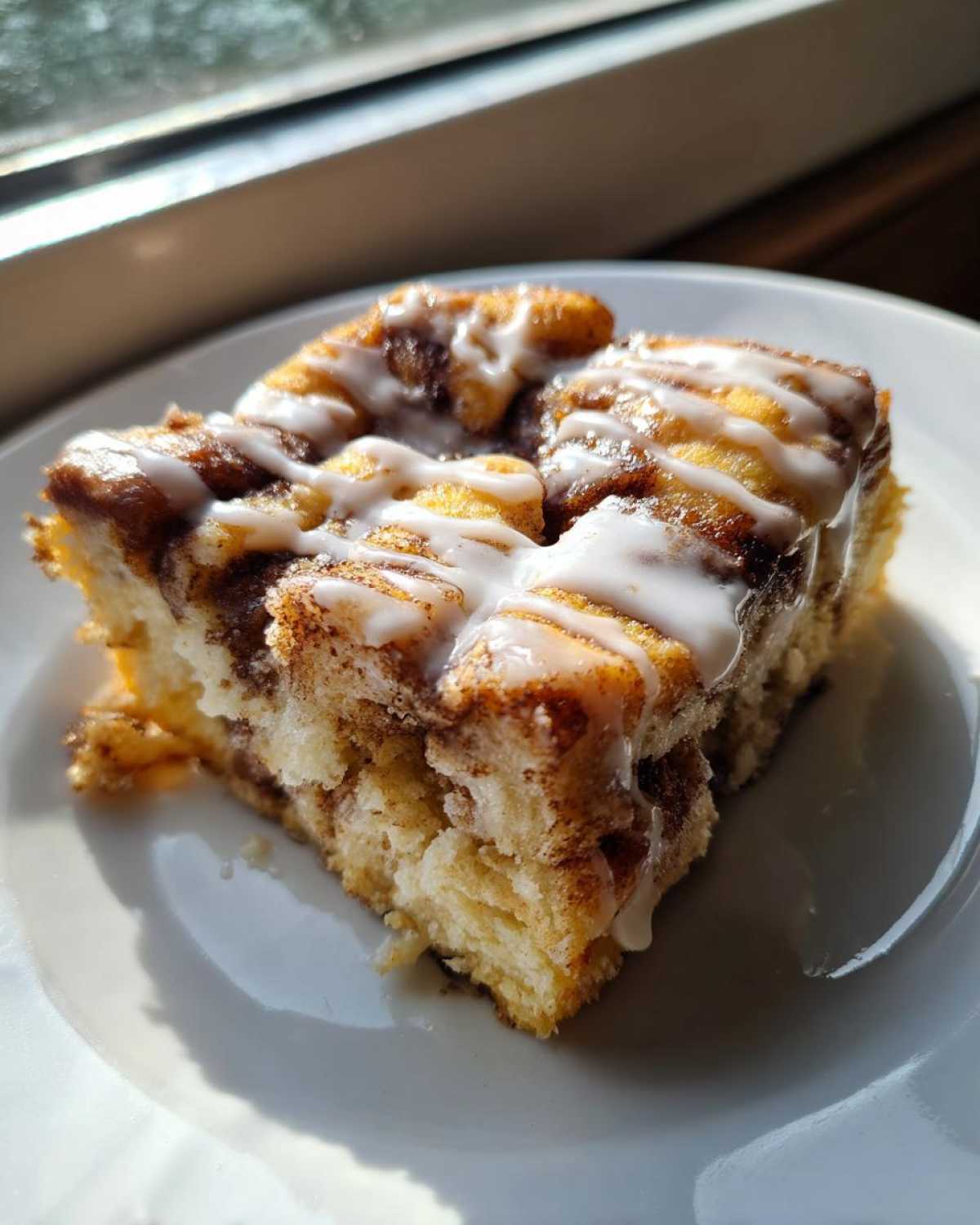 A close-up of a square slice of Easy Sheet Pan Cinnamon Roll Pancake Bake drizzled with white icing on a white plate.