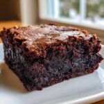 A close-up shot of a rich, fudgy brownie square with a signature crinkly top, resting on a white plate.
