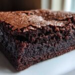 A close-up, mouth-watering shot of a single, dense, fudgy Brownie square with a shiny, cracked top.