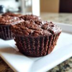 A close-up, appetizing shot of a fudgy brownie muffin sitting on a white square plate, with more brownie muffins blurred in the background.