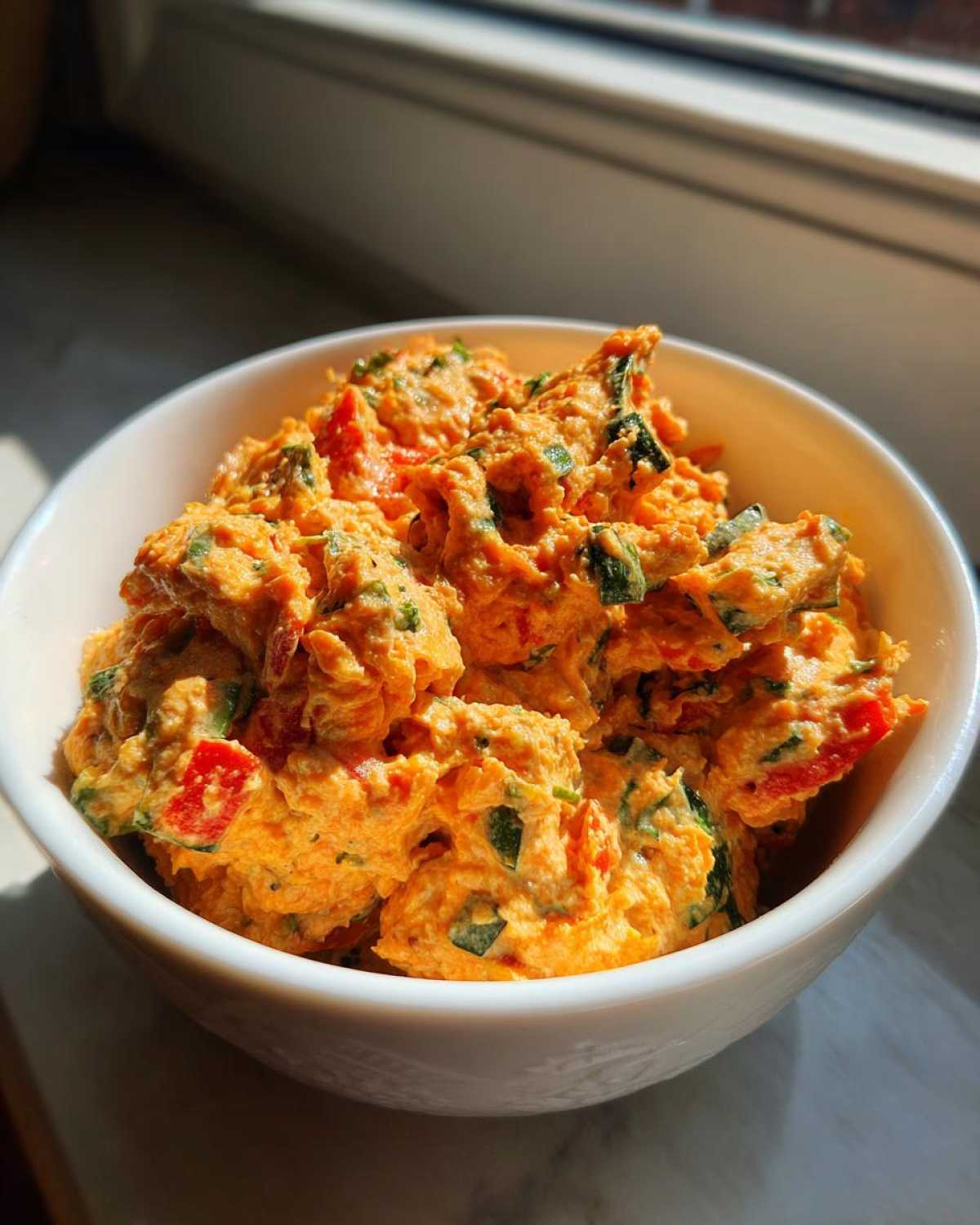 A close-up of a vibrant orange Garden Vegetable Spread mixed with green herbs and red peppers in a white bowl.