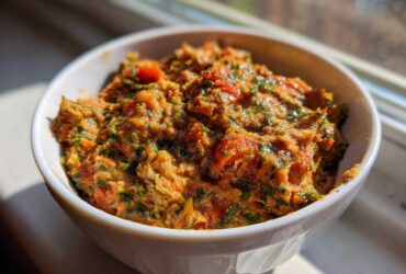 A white bowl filled with a chunky, orange-hued Garden Vegetable Spread mixed with visible green herbs and red tomato pieces.