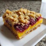A single square of Gluten-Free Lemon Berry Crumble Bars featuring a thick crust, bright yellow lemon filling, dark berry layer, and oat crumble topping.