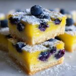 Two stacked squares of bright yellow Gluten-Free Lemon Blueberry Layer Bars, topped with fresh blueberries and powdered sugar.