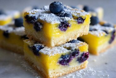 Two stacked squares of bright yellow Gluten-Free Lemon Blueberry Layer Bars, topped with fresh blueberries and powdered sugar.