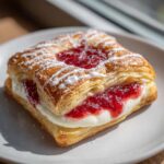 A close-up of a flaky Gluten-Free Strawberry Cream Cheese Danish filled with cream and jam, drizzled with icing.