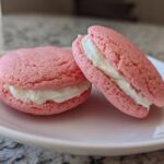 Close-up of two pink Gluten-Free Strawberry Lemon Whoopie Pies filled with white cream, resting on a light pink plate.