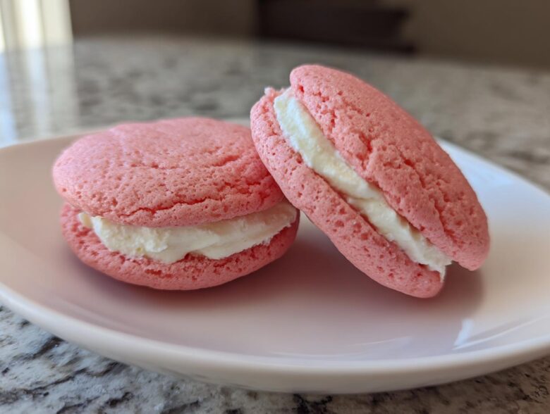 Close-up of two pink Gluten-Free Strawberry Lemon Whoopie Pies filled with white cream, resting on a light pink plate.