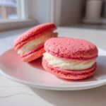 Two bright pink Gluten-Free Strawberry Lemon Whoopie Pies with creamy white filling stacked on a white plate.