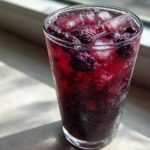 A tall, sweating glass filled with vibrant Iced Blackberry Sage Tea, ice cubes, and whole blackberries.