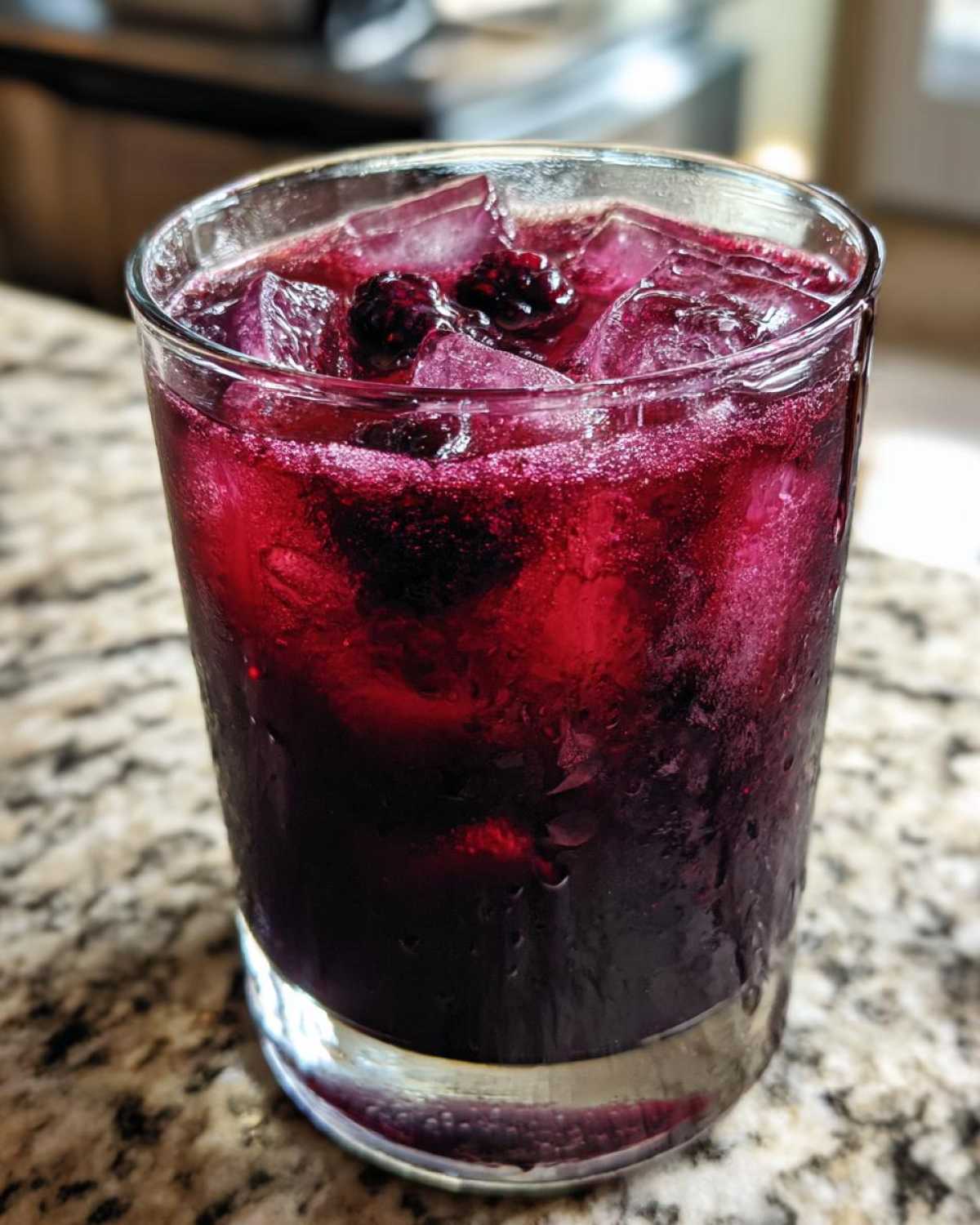 Close-up of a tall glass filled with vibrant, deep purple Iced Blackberry Sage Tea over ice, garnished with blackberries.