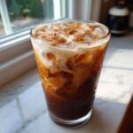A tall glass filled with Iced Brown Sugar Cinnamon Cold Brew, topped with foam and sprinkled cinnamon, showing condensation.