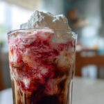 Close-up of a tall glass of Iced Cherry Vanilla Cold Brew swirling with red syrup and topped with whipped cream.