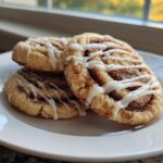 Three delicious Cinnamon Roll Cookies, dusted with cinnamon sugar and drizzled with white icing, stacked on a white plate.