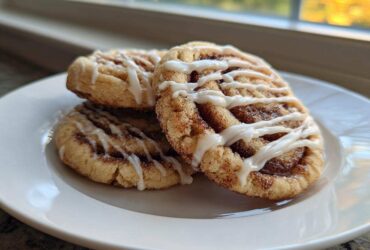 Three delicious Cinnamon Roll Cookies, dusted with cinnamon sugar and drizzled with white icing, stacked on a white plate.