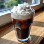 A tall glass of Iced Coconut Mocha with Cold Foam topped with whipped cream sitting on a sunny wooden surface.