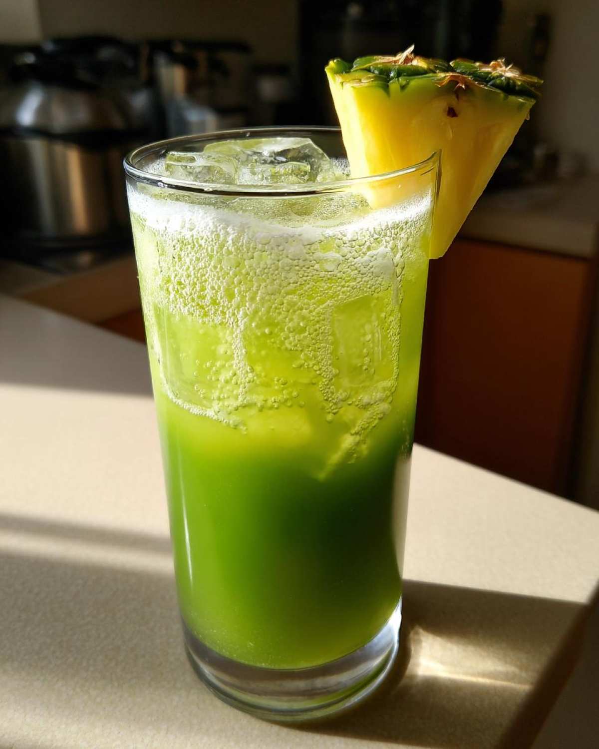 A tall glass filled with a vibrant green Iced Pineapple Matcha Spritz, topped with ice and garnished with a fresh pineapple wedge.