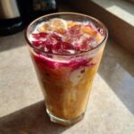 Close-up of a tall glass filled with Iced Raspberry Vanilla Chai, showing layers of orange tea, white cream, and bright magenta raspberry swirl over ice.