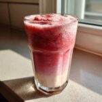 A tall glass filled with a layered Iced Vanilla Strawberry Protein Shake, showing a pink/red frozen top layer and a lighter vanilla base.