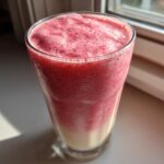 A tall glass filled with a layered Iced Vanilla Strawberry Protein Shake, showing a creamy bottom layer and a frothy pink top layer.
