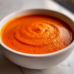 A close-up of a white bowl filled with vibrant orange Instant Pot Creamy Red Pepper Tomato Soup, showing a smooth texture.