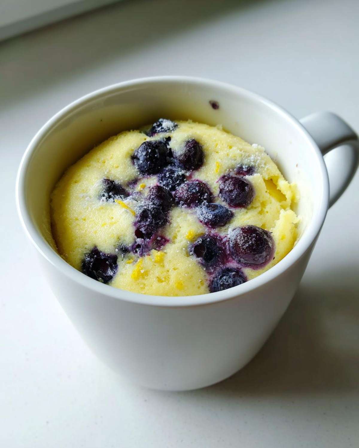 Close-up of a bright yellow Keto Blueberry Lemon Mug Cake topped with whole blueberries and powdered sweetener.