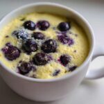 A close-up of a freshly baked Keto Blueberry Lemon Mug Cake, topped with whole blueberries and a dusting of sweetener.