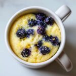 A bright yellow Keto Blueberry Lemon Mug Cake topped with fresh blueberries and lemon zest, served in a white mug.