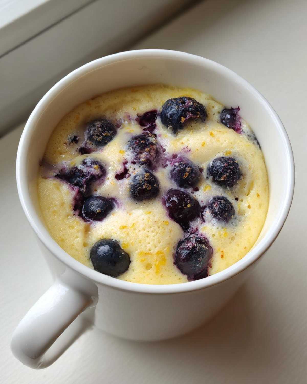 Close-up of a freshly baked Keto Blueberry Lemon Mug Cake, showing blueberries baked into the yellow cake batter inside a white mug.