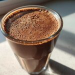 Close-up of a rich, dark brown Keto Creamy Chocolate Peanut Butter Shake with a layer of fine foam on top, sitting by a window.