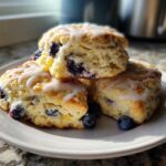 Three fluffy Lemon Blueberry Scones stacked on a white plate, drizzled with white glaze and garnished with fresh blueberries.