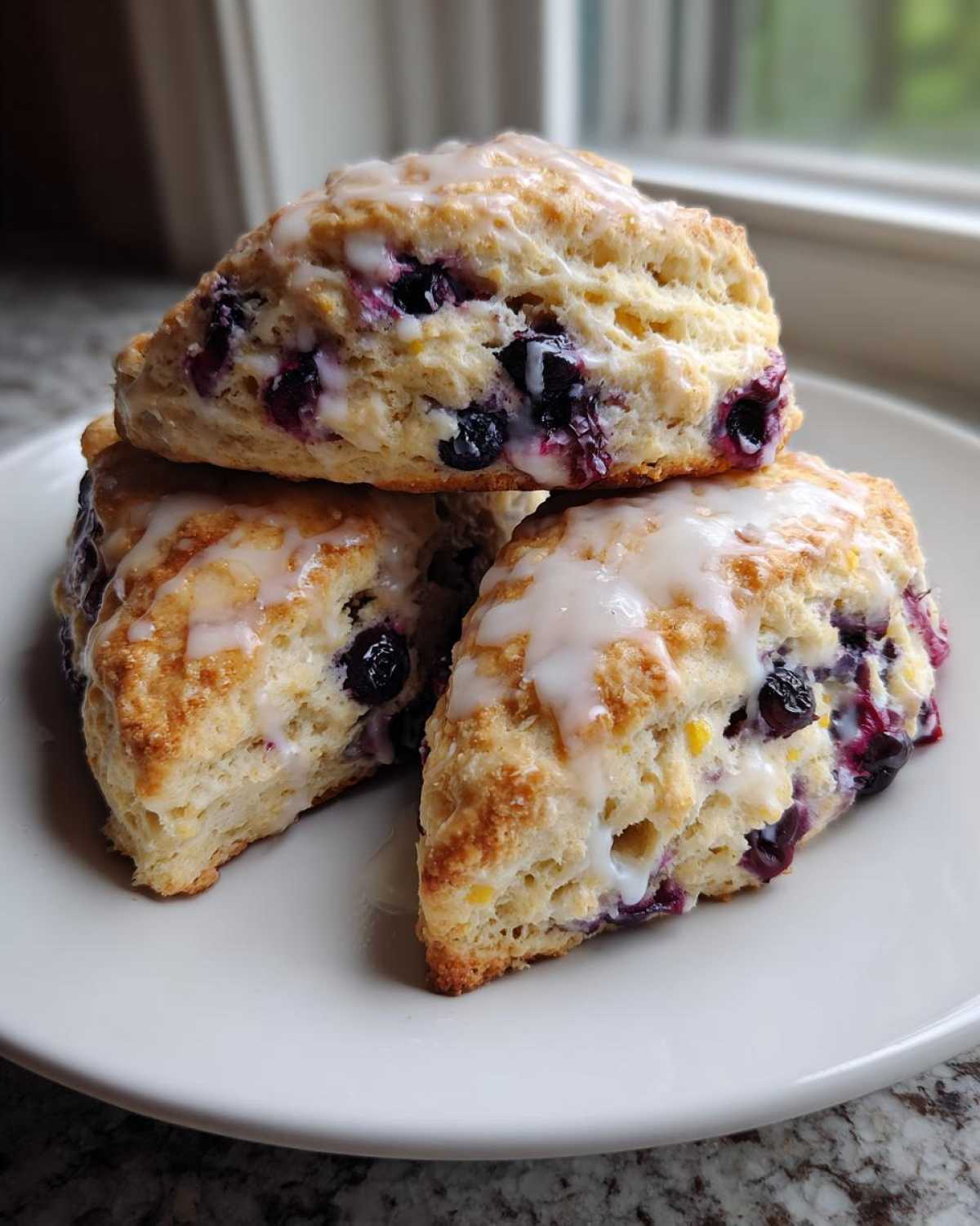 Three freshly baked Lemon Blueberry Scones stacked on a white plate, drizzled with sweet white glaze.