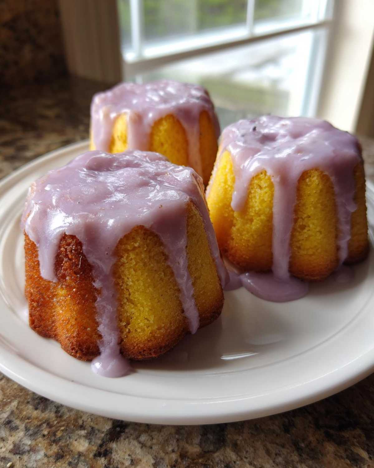 Three bright yellow Mini Lemon Cakes with Lavender Glaze drizzled over the tops, sitting on a white plate.