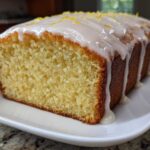 Close-up of a freshly baked, moist Lemon Loaf with thick white glaze dripping down the sides and topped with lemon zest.