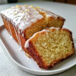 A moist Lemon Loaf cake, partially sliced, topped with white glaze icing and lemon zest.