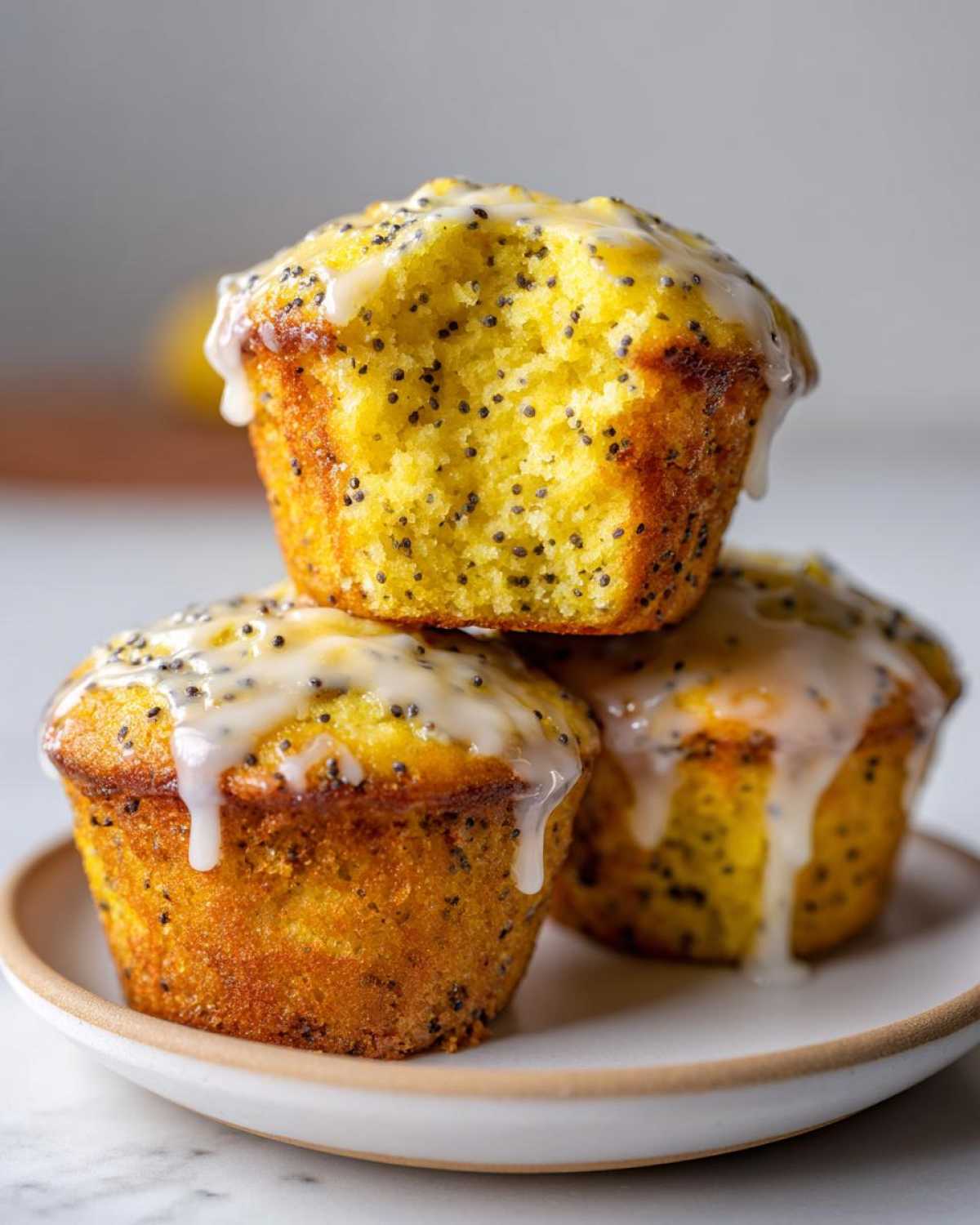 Three moist Lemon Poppy Seed Muffins stacked on a plate, drizzled with a thick white lemon glaze.