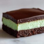 A single serving of No-Bake Chocolate Mint Icebox Bars showing distinct layers: a dark chocolate crust, a bright mint green filling, and a smooth chocolate ganache topping.