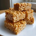 A stack of gooey, golden No-Bake Peanut Butter Rice Cereal Bars sitting on a white plate.