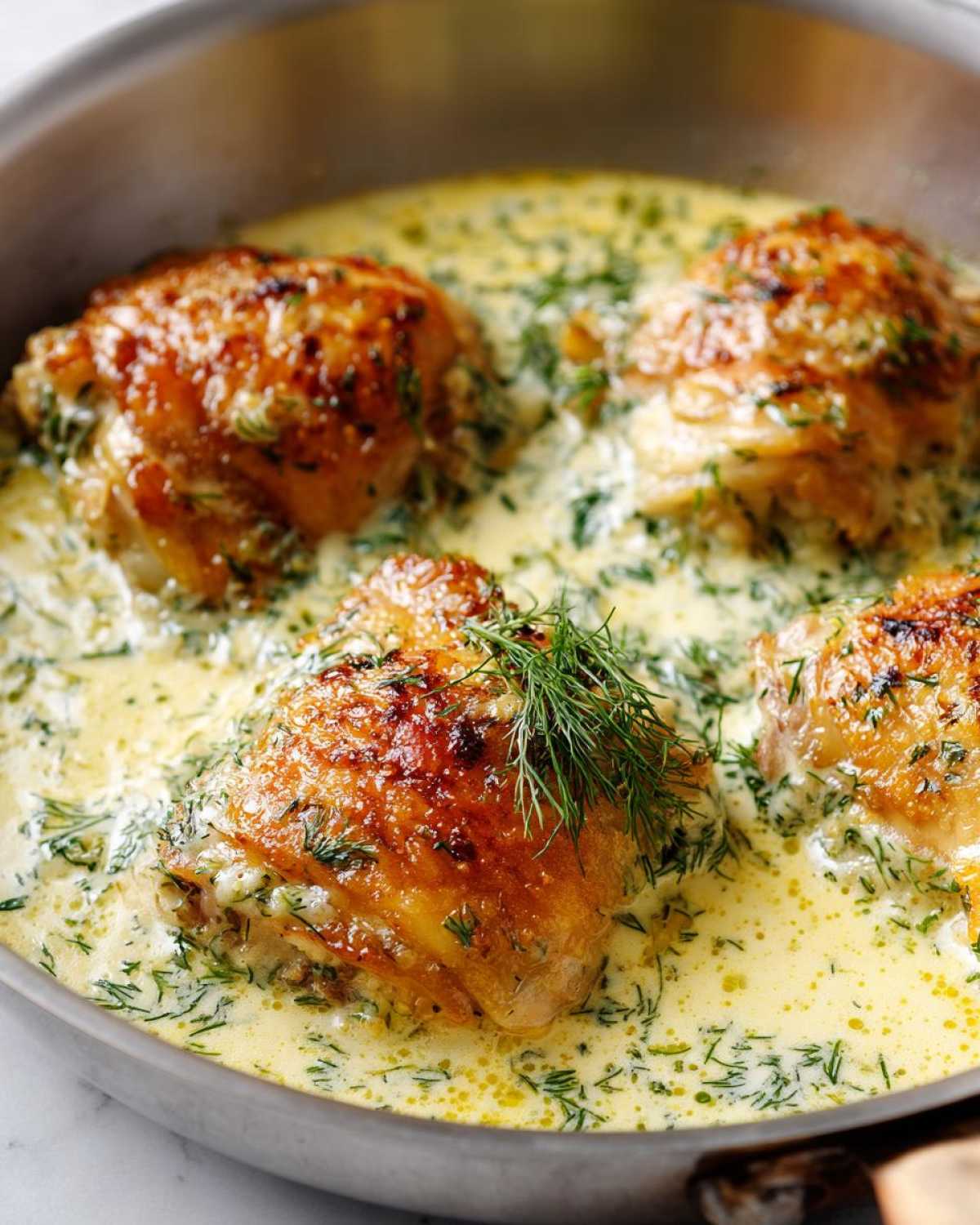 Four golden-brown chicken thighs simmering in a creamy lemon dill sauce filled with fresh dill in a stainless steel pan.