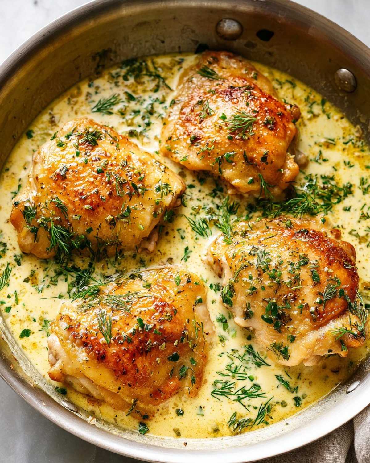 Four golden brown chicken thighs simmering in a creamy lemon dill sauce, garnished with fresh dill.