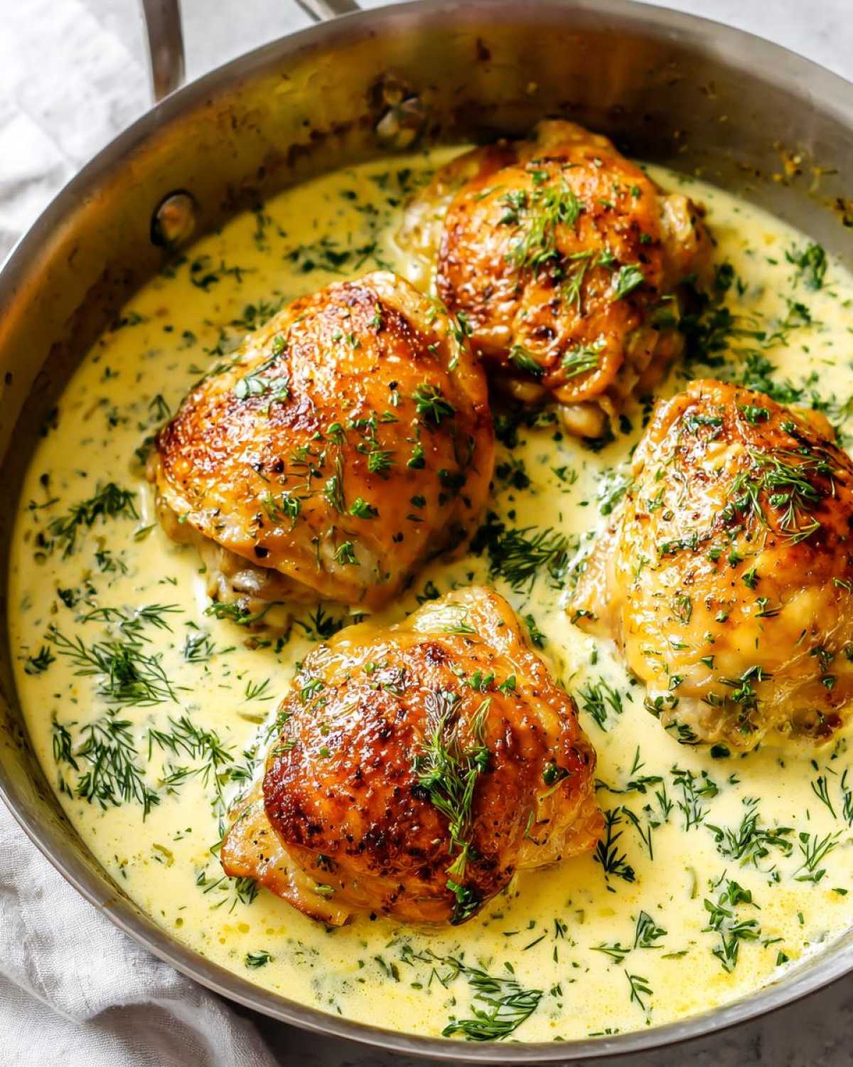Four golden-brown chicken thighs simmering in a creamy lemon dill sauce, garnished with fresh dill.