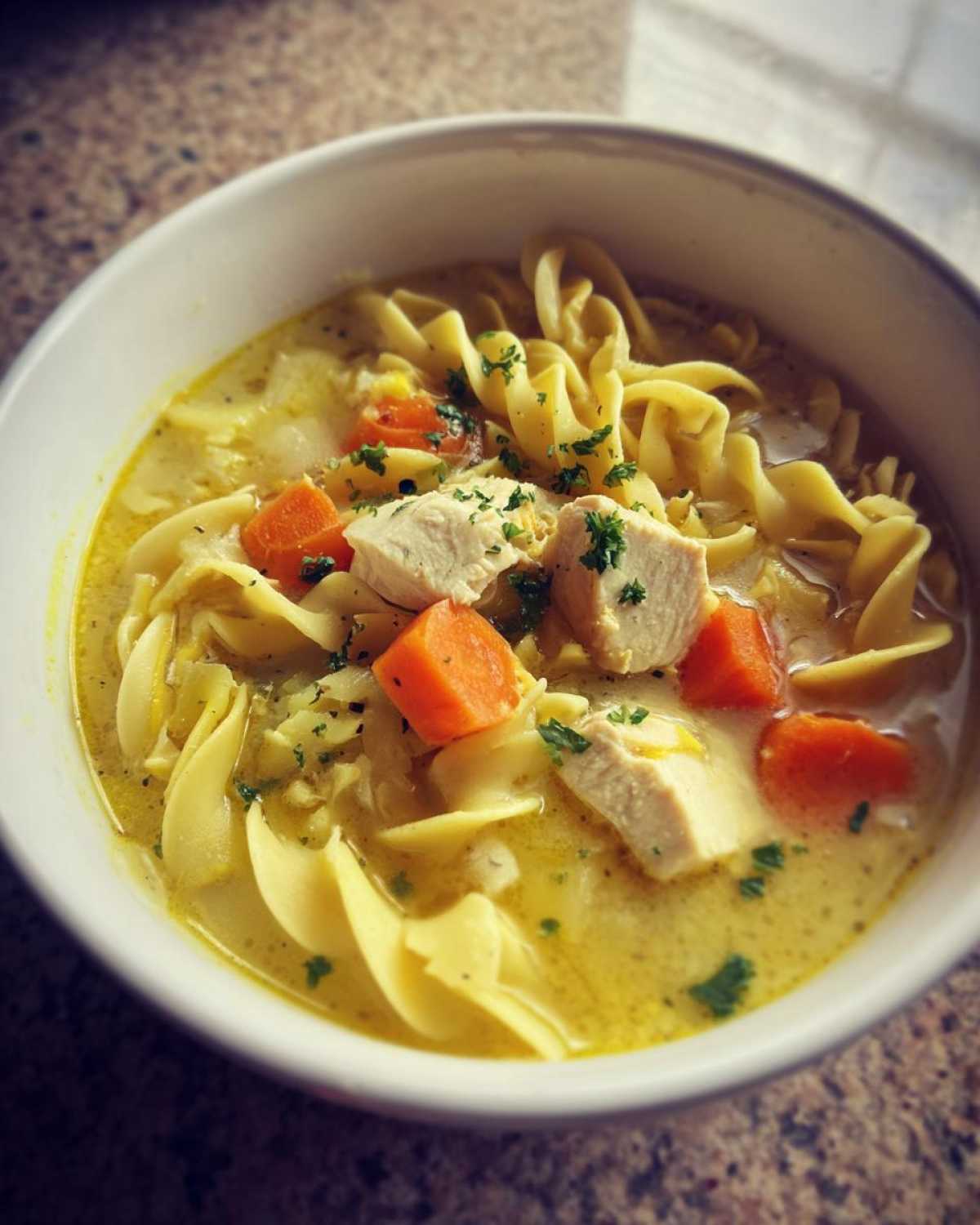 A close-up view of a white bowl filled with One-Pot Creamy Lemon Chicken Noodle Soup, featuring egg noodles, carrots, and chicken chunks.
