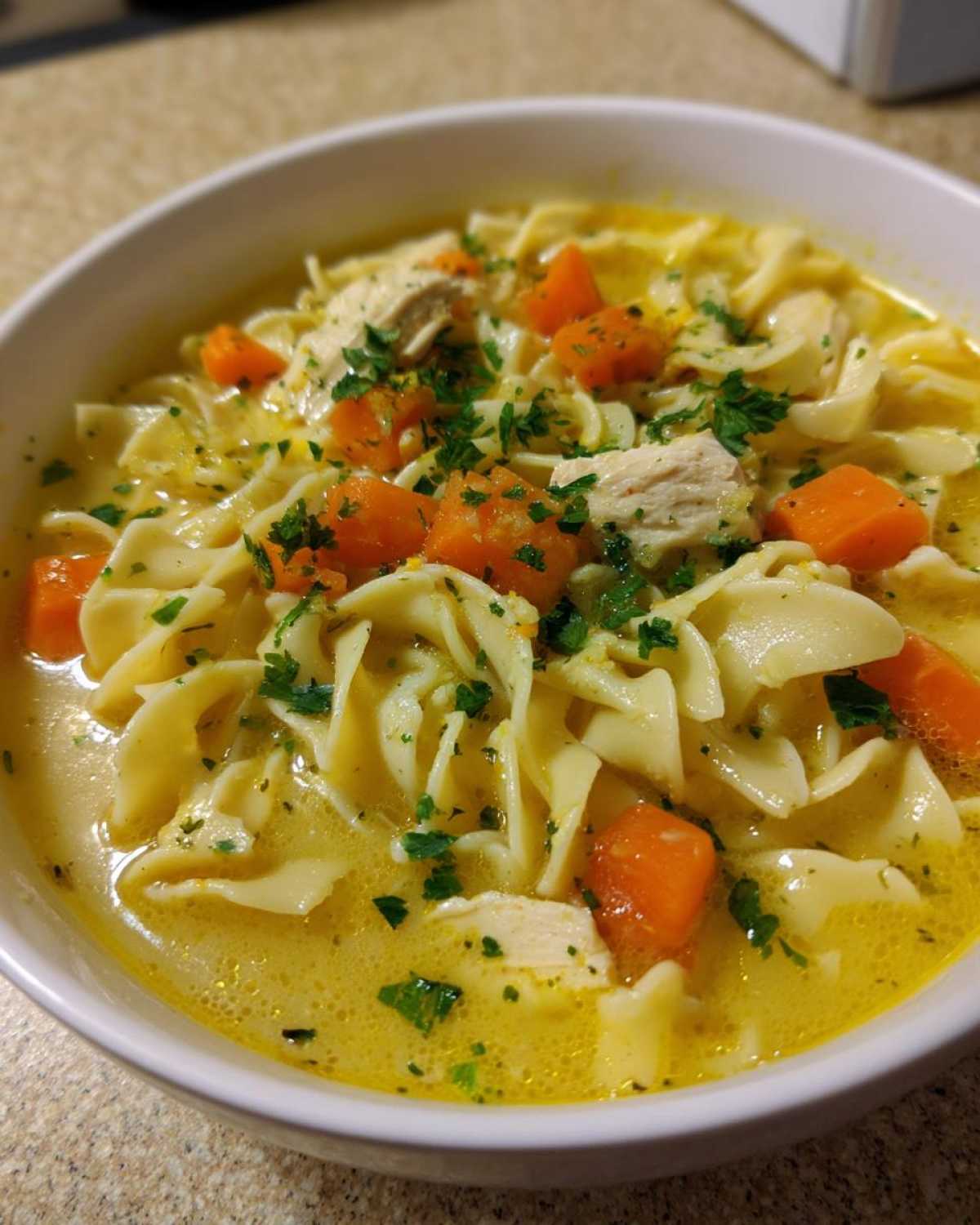 A close-up view of a white bowl filled with One-Pot Creamy Lemon Chicken Noodle Soup, featuring egg noodles, carrots, and chicken.