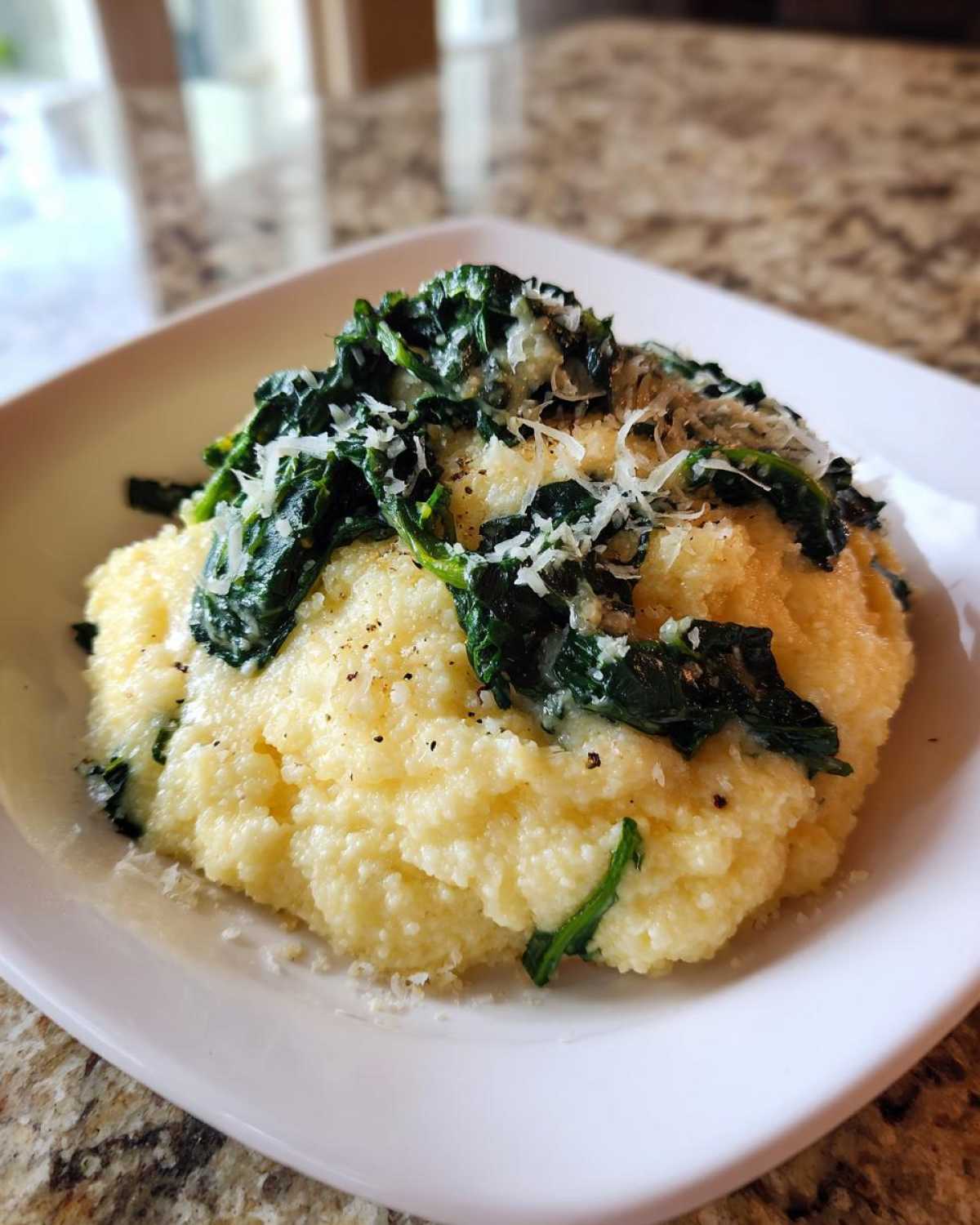 A close-up of One-Pot Creamy Spinach Parmesan Polenta served on a white square plate, topped with wilted spinach and grated cheese.