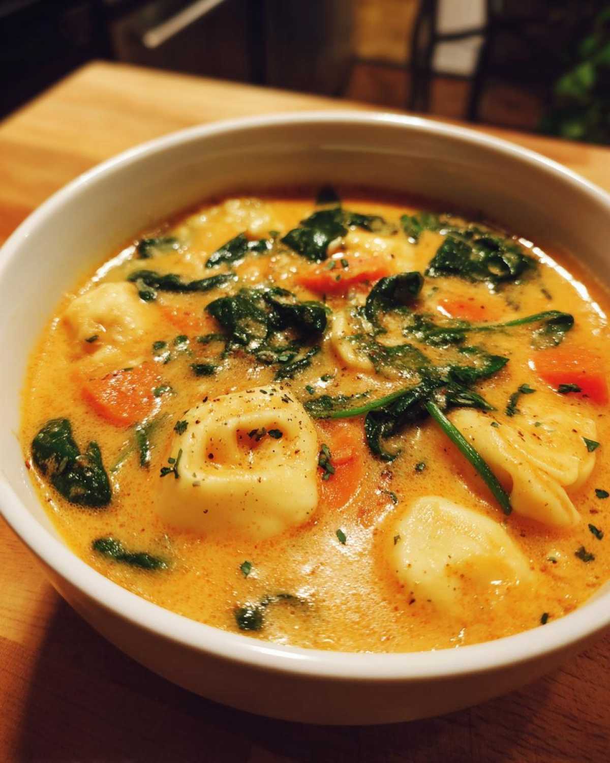 A close-up of a white bowl filled with One-Pot Creamy Tortellini Minestrone, featuring tortellini, spinach, and carrots in a rich orange broth.