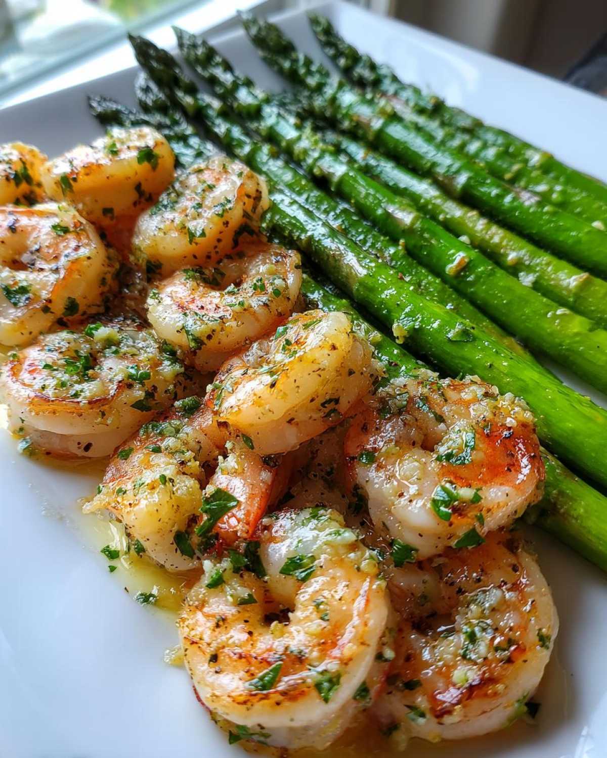 Close-up of Sheet Pan Crispy Garlic Butter Shrimp and Asparagus glistening with butter sauce on a white plate.