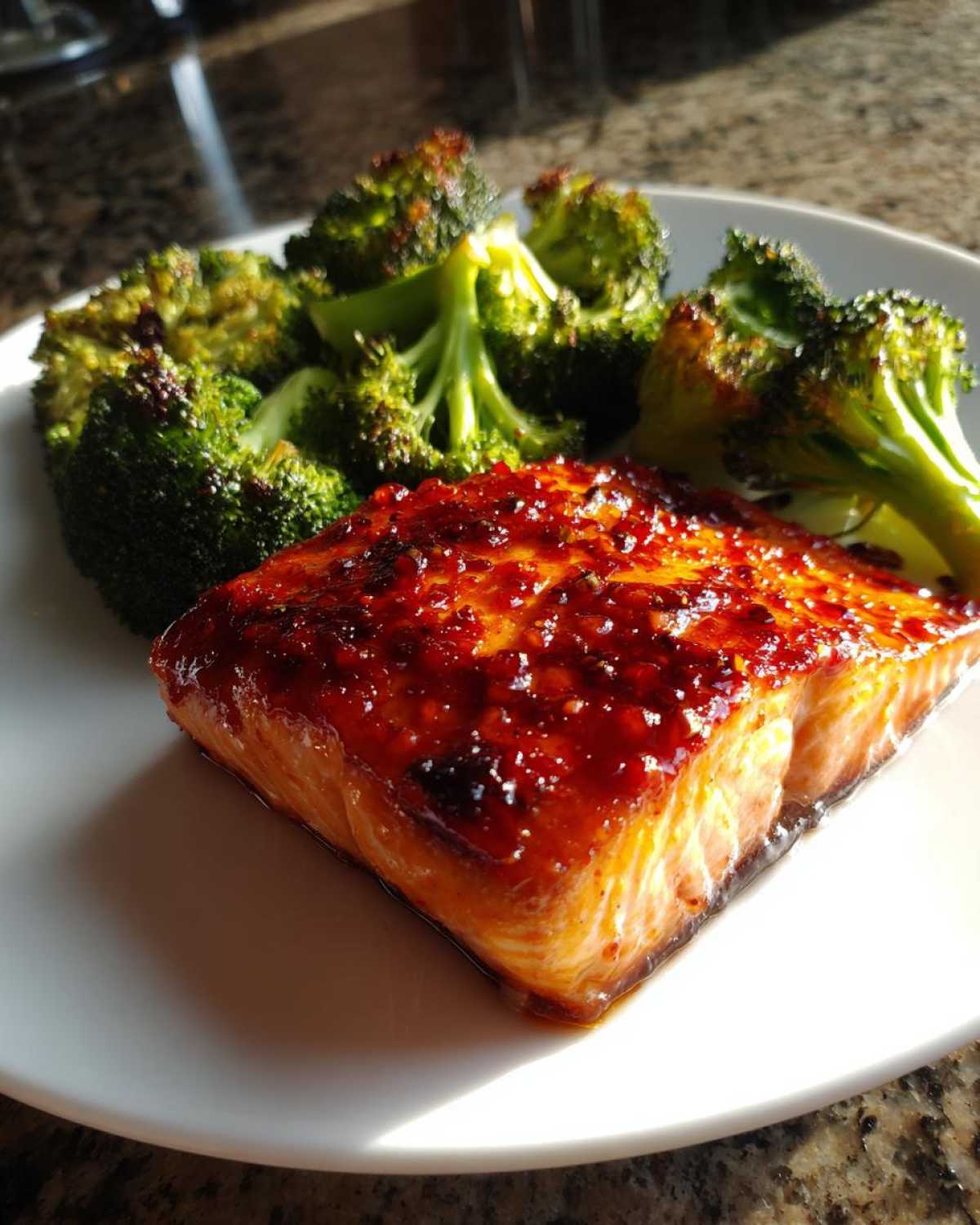 A perfectly cooked fillet of Sheet Pan Honey Harissa Salmon with a glossy red glaze, served next to roasted broccoli florets on a white plate.