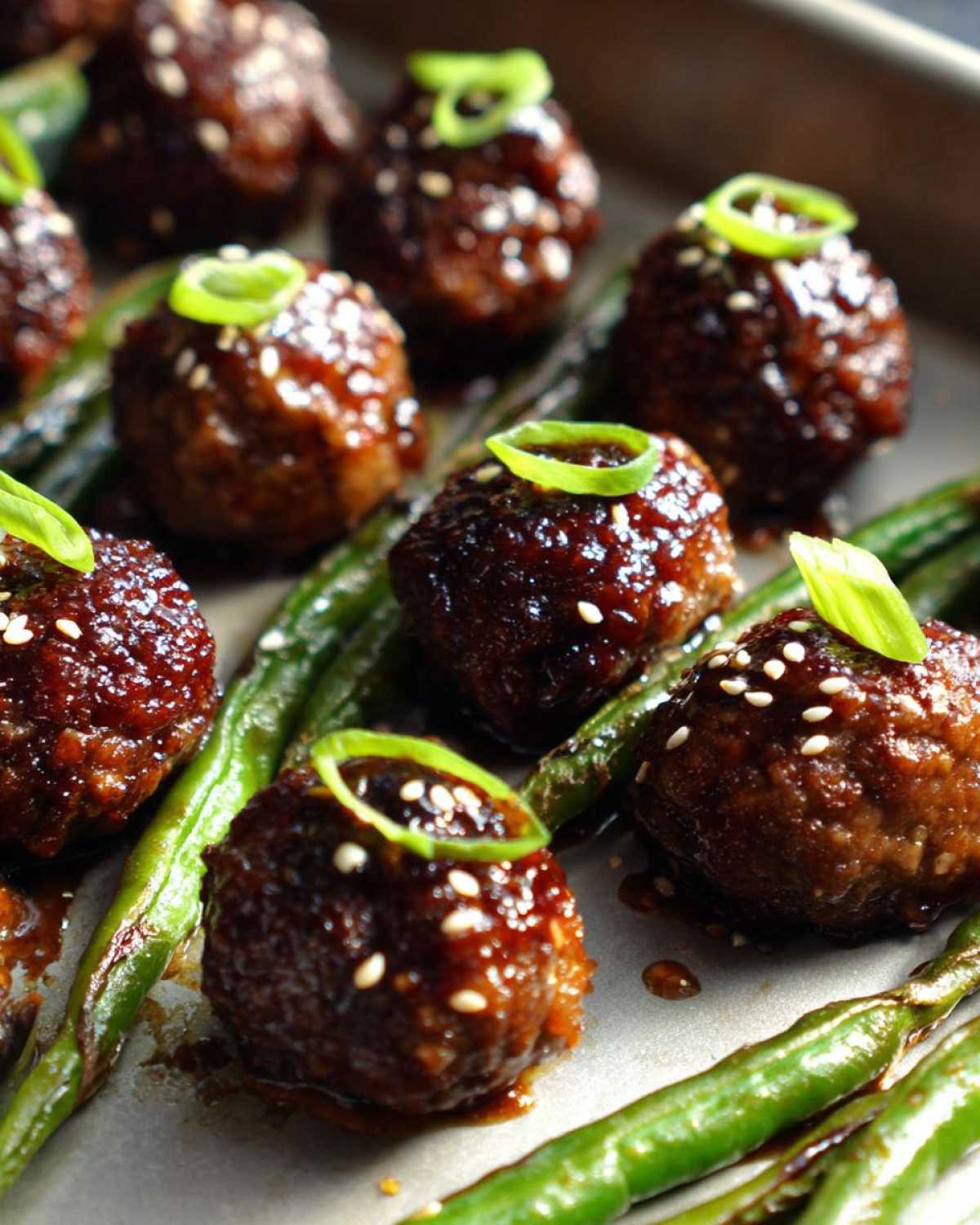 Close-up of Sheet Pan Sticky Sesame Meatballs glazed with sauce, sprinkled with sesame seeds, and garnished with green onion rings, resting on roasted snap peas.