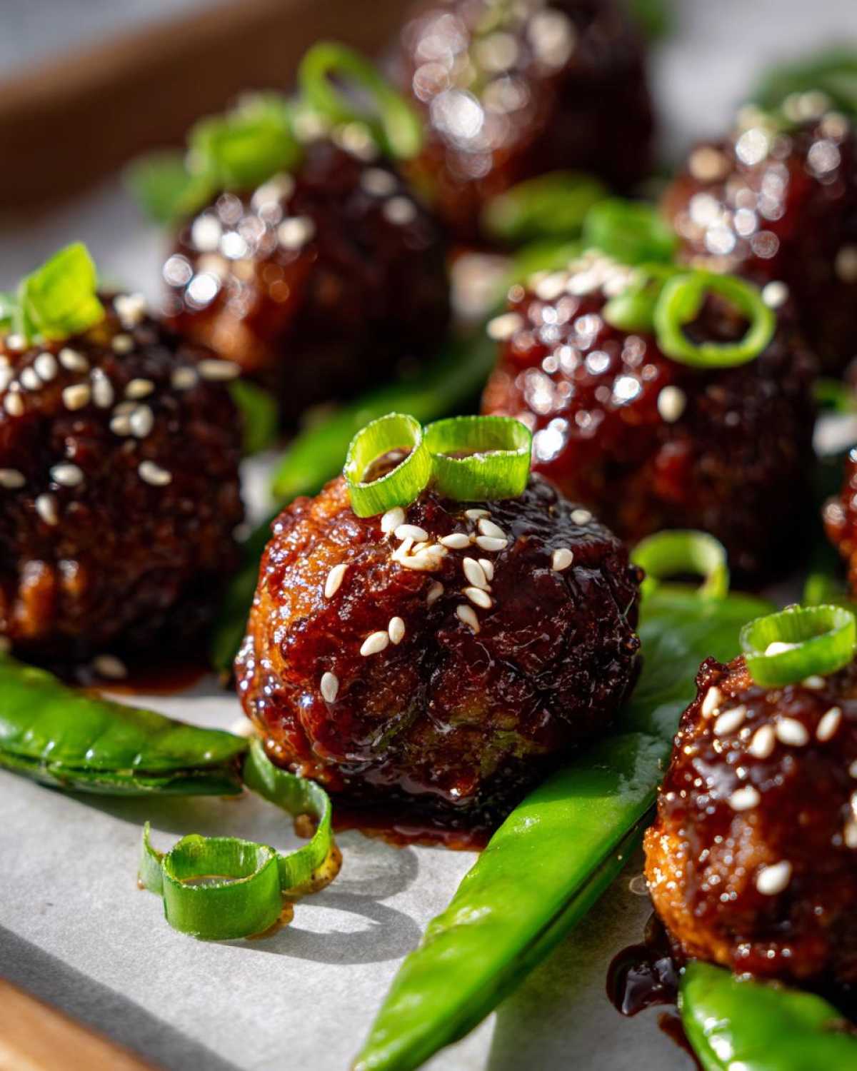 Close-up of glazed Sheet Pan Sticky Sesame Meatballs garnished with sesame seeds and green onions atop bright green snap peas.